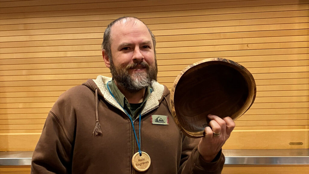 Nate shows a wooden bowl with a natural edge that he turned on a lathe. He also wears a name badge.