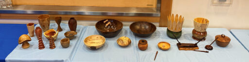 A table with dozens of woodturning projects, mostly bowls, platters, and vessels. Also, there are woodturned pens, trees, a mushroom, and two plant pots.