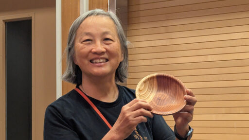 Alison Lee smiles warmly while holding up a hand-turned wooden bowl at the West Bay Woodturners meeting in May 2026. She turned the bowl from redwood. The bowl features distinctive light-colored sapwood on the left and deep reddish-brown heartwood on the right.
