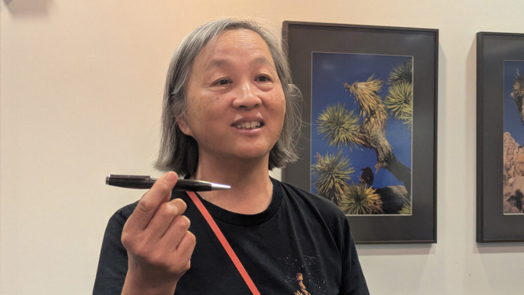 Alison Lee smiles while holding a dark, turned pen horizontally in her right hand. In the background, framed photographs of Joshua trees hang on a white wall at the West Bay Woodturners meeting.