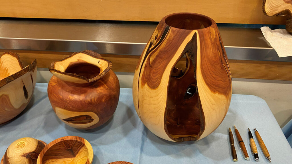 Three turned wooden vessels, and four pens made from Juniper wood are displayed on a blue tablecloth. The largest object is a tall, sculptural vase on the right, featuring a large open cutout that reveals dark wood grain against the lighter exterior, turned by Bob Bley. Next to it is a smaller, rounder pot with a wide, irregular natural rim. Partial views of another vessel and small bowls are visible on the left, while four matching turned pens lie vertically on the right.