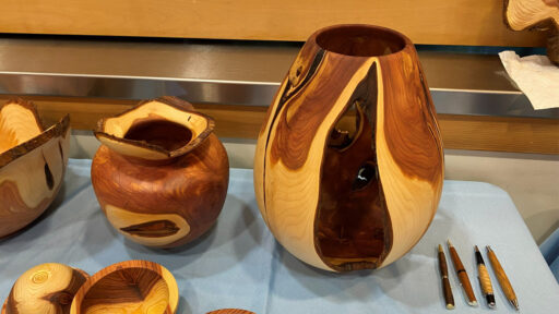 Three turned wooden vessels, and four pens made from Juniper wood are displayed on a blue tablecloth. The largest object is a tall, sculptural vase on the right, featuring a large open cutout that reveals dark wood grain against the lighter exterior, turned by Bob Bley. Next to it is a smaller, rounder pot with a wide, irregular natural rim. Partial views of another vessel and small bowls are visible on the left, while four matching turned pens lie vertically on the right.