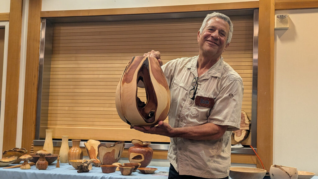 Bob, wearing a name tag, smiles as he holds up a large, bulbous hollowform turned from Juniper with a distinctive opening and contrasting light and dark wood grain. He stands behind a table displaying a variety of other wooden turning projects, including bowls and vases, at a West Bay Woodturners meeting.