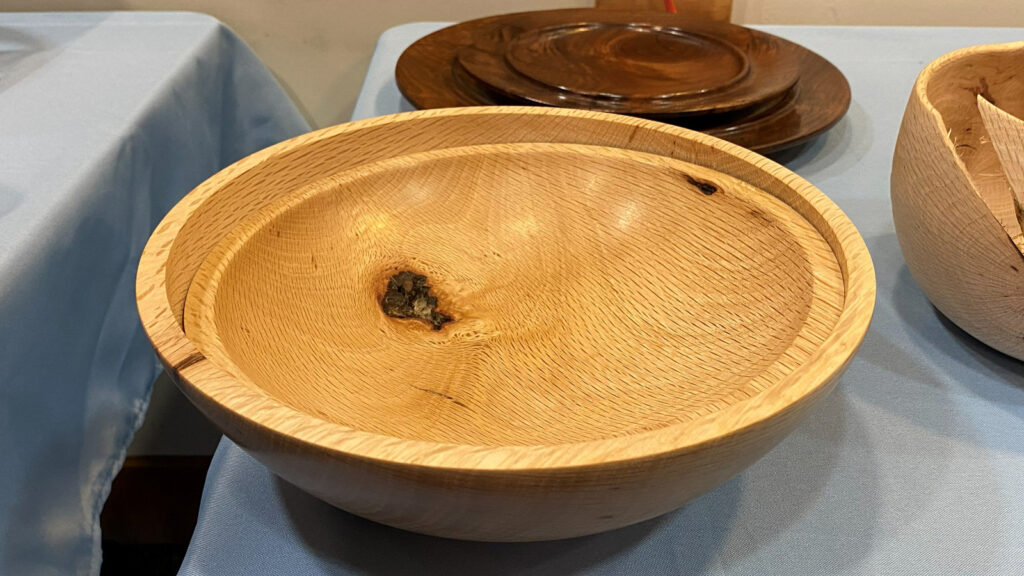 Large, nested, wooden bowls turned from light-colored oak, featuring a prominent swirling grain pattern and a dark knot near the center. It is displayed on a table covered with a light blue cloth at a woodturning club meeting.