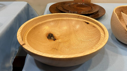 Large, nested, wooden bowls turned from light-colored oak, featuring a prominent swirling grain pattern and a dark knot near the center. It is displayed on a table covered with a light blue cloth at a woodturning club meeting.
