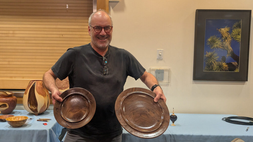 Daniel holds up two large, dark wood turned platters, one in each hand, at a West Bay Woodturners meeting. He stands in front of a table covered with a light blue cloth that displays other woodturning projects, including a large two-toned vase and smaller wooden bowls. A framed picture of a Joshua tree hangs on the wall behind him.