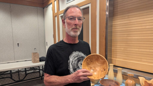 Dean Caudle holds a light-colored turned cherry bowl at the West Bay Woodturners meeting. He wears a black t-shirt with a wood ring design. Other turned wood projects, including vases and a dark bowl, are displayed on a table in the foreground.