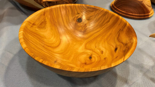A close-up, high-angle shot of a turned cherry wood bowl by Dean Caudle, featuring a lustrous, golden-orange finish with prominent swirling grain patterns and several dark knots. The bowl sits on a light blue tablecloth, displayed as part of the recent woodturning projects at the West Bay Woodturners meeting, with other wooden pieces partially visible in the background.