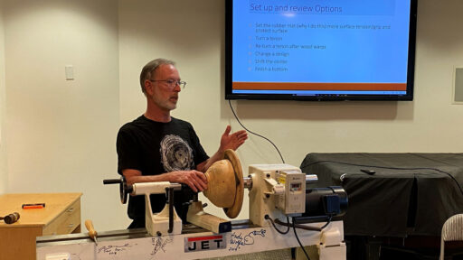 Dean Caudle stands behind a wood lathe, gesturing with his left hand as if explaining a technique during a demonstration. A light-colored wooden bowl is mounted on the lathe. Behind him, a large screen displays a presentation slide titled "Set up and review Options" with a numbered list of instructions such as "Set the rubber mat" and "Turn a tenon." Signatures of famous turners are written on the lathe. The picture was taken for the woodturners newsletter.
