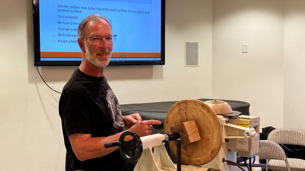 Dean Caudle stands beside a wood lathe, gesturing toward a square block of wood mounted on a large wooden faceplate. He is wearing a black t-shirt and smiling. Behind him, a large flat-screen monitor displays a blue presentation slide with a numbered list of instructions; visible text includes "1. Set the rubber mat (why I do this), more surface tension/grip and protect surface" and "2. Turn a tenon." The setting is a woodturning workshop with Dean Caudle giving a woodturning demonstration.