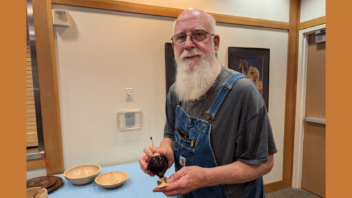 Harvey Klein holds a dark, polished wooden turned box while standing at a West Bay Woodturners meeting. He is positioned behind a table covered with a blue cloth, displaying several light-colored wooden bowls and a dark plate. In the background, a framed picture of Joshua trees on a wall with dark wood trim.