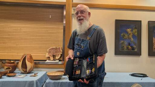 Harvey Klein, a local woodturner, smiles as he holds open a black case displaying a collection of colorful, hand-turned wooden pens. In the background, a table displays other woodturning projects, including wooden bowls and vessels, at a West Bay Woodturners club meeting. The picture was taken for the West Bay Woodturners newsletter.