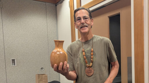 Kelly holds up a polished, golden-brown wooden vase (hollowform) in his right hand. He is wearing a green t-shirt and a large necklace of wooden beads with a pendant engraved with the name "Kelly Smith." He is presenting his woodturning project at a West Bay Woodturners club meeting.