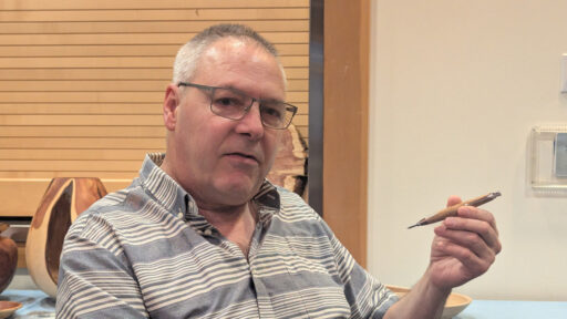Kevin Lee holds up a turned wooden pencil during a West Bay Woodturners meeting. In the background, wooden bowls sit on a table against a slatted wood wall.