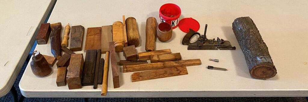 An overhead shot of a white table displaying an assortment of woodturning materials and tools. The collection includes various rectangular blocks of timber in shades of dark brown and light tan, turned wooden spindles and handles, and rough logs. To the right, a large log with bark sits next to a metal hand plane, a metal compass, a small wooden cup, and a red plastic bucket. The arrangement shows supplies for a woodworking project.