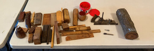 An overhead shot of a white table displaying an assortment of woodturning materials and tools. The collection includes various rectangular blocks of timber in shades of dark brown and light tan, turned wooden spindles and handles, and rough logs. To the right, a large log with bark sits next to a metal hand plane, a metal compass, a small wooden cup, and a red plastic bucket. The arrangement shows supplies for a woodworking project.