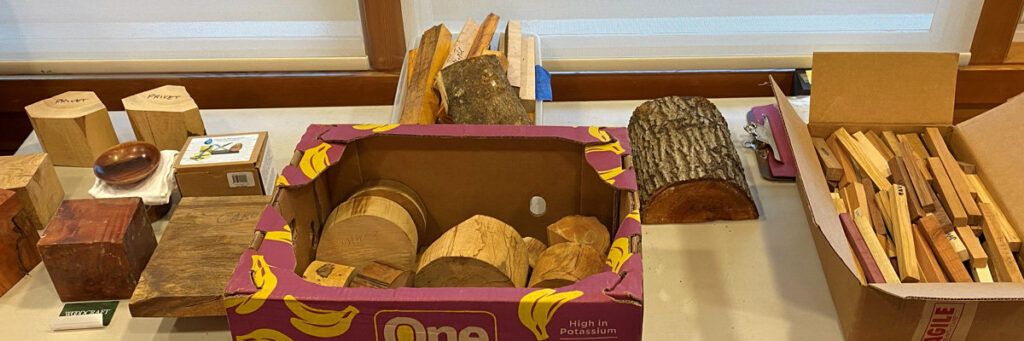 A tabletop display featuring various woodworking projects and raw materials. On the left are several round and hexagonal wooden blocks and a flat board with the word "Carob" written on it. In the center, a purple cardboard box is filled with round wood slices (cookies). Behind this box are raw lumber scraps and a large segment of a log. To the right, a large open cardboard box is packed with numerous small, wooden pen blanks.