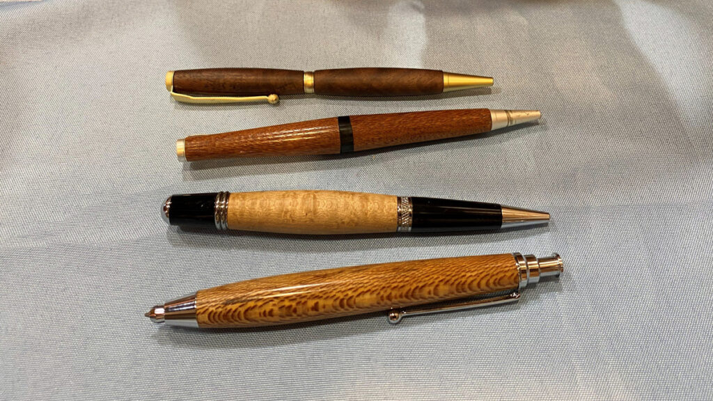 Four hand-turned wooden pens arranged horizontally on a light blue, textured fabric background.