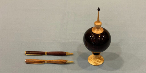 Two turned wood pens lie horizontally on a light grey surface next to a matching lidded container. To the right stands a spherical dark-wood jar with a light-wood base and a lid topped with a slender, pointed finial.