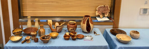 An eye-level, wide shot, taken for the West Bay Woodturners newsletter, captures a display of woodturning projects arranged on tables covered with light blue tablecloths. The collection features a diverse array of turned wooden vessels, including tall slender vases, natural-edge bowls, lidded jars, and serving platters. The pieces exhibit a wide range of wood grains and colors, from pale sapwood to dark heartwood, with some items featuring dramatic live edges or split designs. Prominent pieces include a tall, large vase with a unique open center and a set of small nesting bowls. On the windowsill in the background, a cross-grain slab of Juniper with a natural edge is resting against a wooden slat blind.