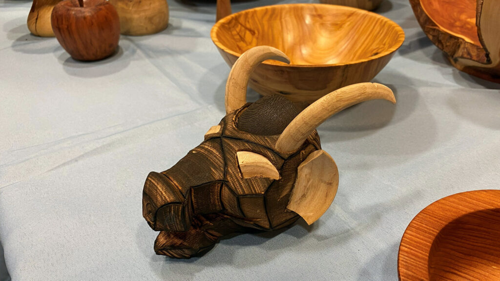 A close-up view of a wooden sculpture depicting a dragon head with a geometric, faceted design. The main body of the head is turned and burned wood with angular segments, while the upward-curving horns, ear, and eye area are made from lighter, natural wood. The sculpture sits on a light blue tablecloth, surrounded by other woodturning projects in the background, including a large wooden bowl and a wooden apple, at a display for the West Bay Woodturners club.