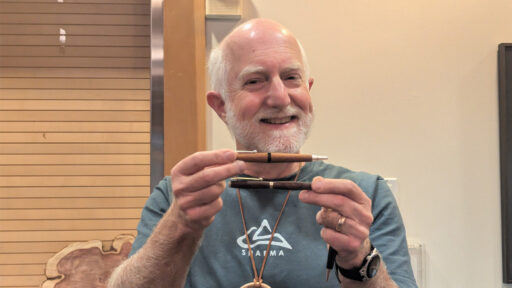 Vic smiles and holds up three handcrafted wooden pens, displaying them horizontally and one vertically in front of his chest. The background features a wall with horizontal slats, consistent with a woodturning club meeting setting.