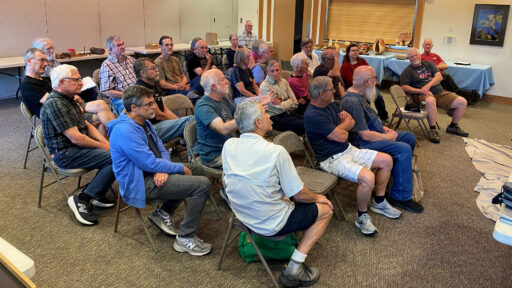 A group of attendees, primarily men with a few women, sit in rows of folding chairs facing right during a woodturning demonstration. In the background, tables display various wooden bowls and vessels.