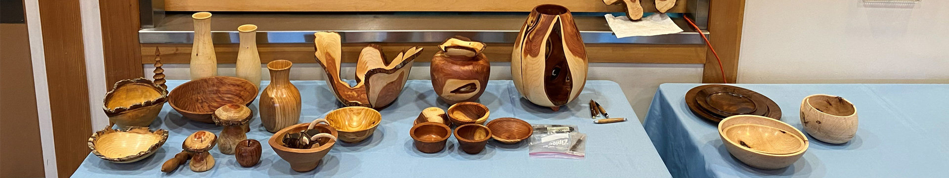 A wide shot of a woodturning show display featuring a variety of turned wooden objects arranged on tables covered with light blue tablecloths. The collection includes live-edge bowls, tall slender vases, a large open sculptural bowl, a lidded jar, and numerous small bowls in various woods and grain patterns. A tall, prominent vase with a split design in contrasting light and dark wood stands near the center. To the right, a separate table section holds stacked dark wooden platters and lighter wooden bowls.