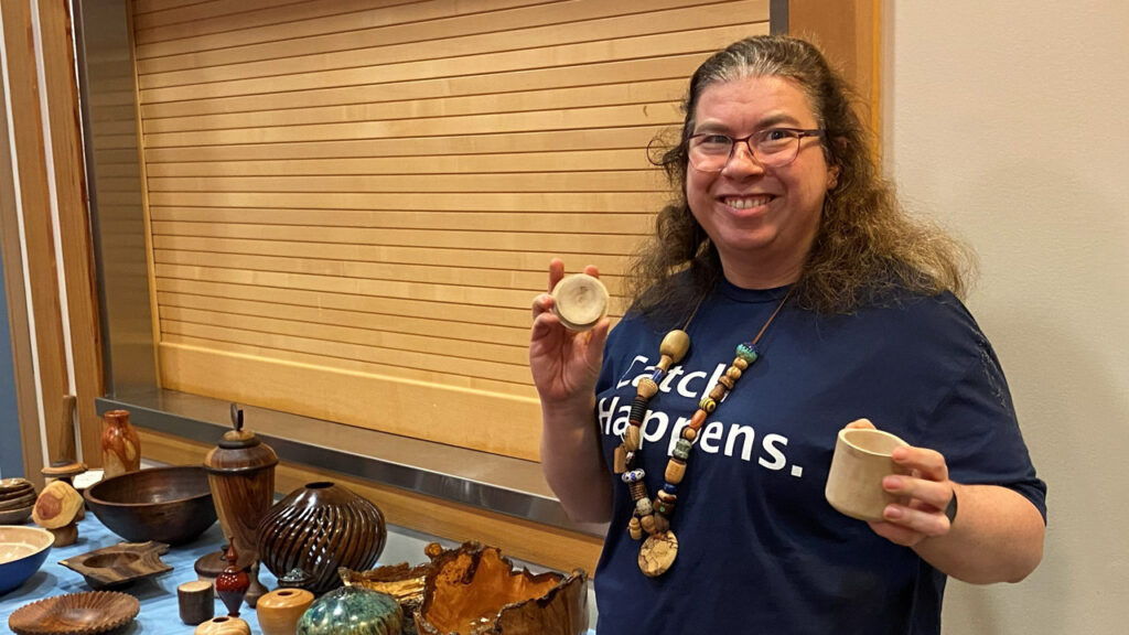 Angela Gunn, a founder of Turned Connections, smiles while displaying a woodturned lidded box from light-colored wood at a West Bay Woodturners meeting. She wears glasses, a large beaded necklace (all beads are unique, and were turned by different artists), and a blue T-shirt that reads "Catch Happens." A table in front of her is covered with an assortment of turned wood items, including dark bowls, ribbed vessels, and spindles, set against a background of horizontal wooden slats.