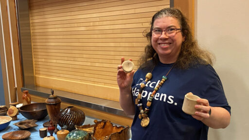 Angela Gunn, a founder of Turned Connections, smiles while displaying a woodturned lidded box from light-colored wood at a West Bay Woodturners meeting. She wears glasses, a large beaded necklace (all beads are unique, and were turned by different artists), and a blue T-shirt that reads "Catch Happens." A table in front of her is covered with an assortment of turned wood items, including dark bowls, ribbed vessels, and spindles, set against a background of horizontal wooden slats.