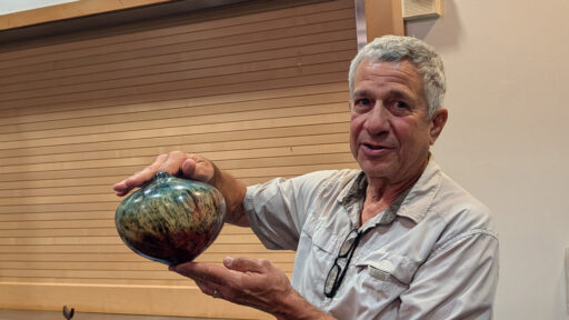Bob Bley holds a glossy, bulbous woodturned vessel. The piece features a swirling finish in teal, green, and dark brown. He is wearing a light beige button-down shirt, with glasses hanging from the collar, and stands in front of a wall of horizontal wooden slats.
