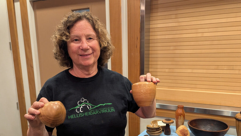 Carole Gilbert smiles while holding two turned wooden hollow forms, one round bowl in her right hand and a taller cup-shaped vessel in her left at a West Bay Woodturners meeting. She is wearing a black T-shirt with a green logo. To her right, a table displays other woodturning projects, including a stack of small bowls, a tall vase-like piece, and a large dark bowl, set against a wall of light horizontal wood paneling.