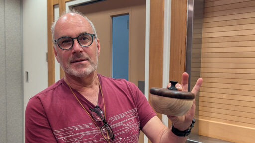 Daniel Saal, a man with glasses and a short grey beard, holds up a small, turned wooden lidded bowl in his left hand. He is wearing a maroon t-shirt with a white circuit-board style pattern and has a pair of reading glasses hanging from the collar of his shirt. The background features light wood paneling and a doorway. He presents his woodturning project at a West Bay Woodturners club meeting.