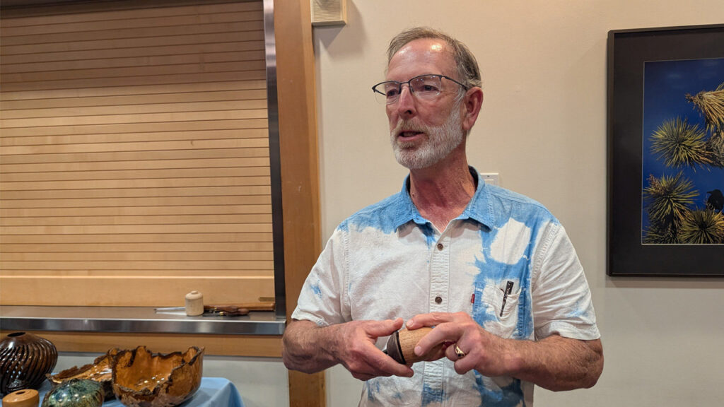A bearded man, Dean Caudle, with gray hair and glasses, wearing a white short-sleeved shirt with blue tie-dye patterns, stands holding a small turned wooden box in the shape of an acorn in his hands. He appears to be speaking or explaining the object. To his left, on a table covered with a light blue cloth, is a display of various woodturning projects, including a dark ribbed vase, a natural-edge bowl, and other small turned items. Behind him on the wall hangs a framed photograph of spiky plants against a blue sky. The setting appears to be an indoor meeting for the West Bay Woodturners club.