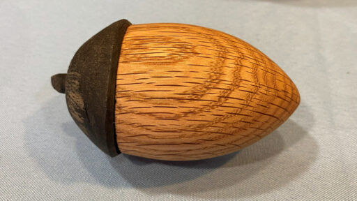 A hand-turned wooden acorn-shaped box resting on a light-blue cloth surface. The main body of the acorn is crafted from light golden-brown wood with prominent, horizontal grain patterns, while the cap is made of a contrasting dark brown wood. A small, dark stem protrudes from the left side where the lid meets the base, indicating it is a lidded container.
