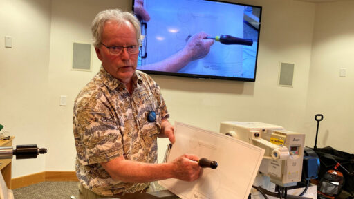 At a West Bay Woodturners meeting, Dennis Lillis demonstrates thread-chasing turning techniques. He stands in the foreground holding a large paper template and a black tool handle, looking toward the audience. Behind him, a wall-mounted television displays a live close-up feed of hands performing the same technique on a similar paper drawing.