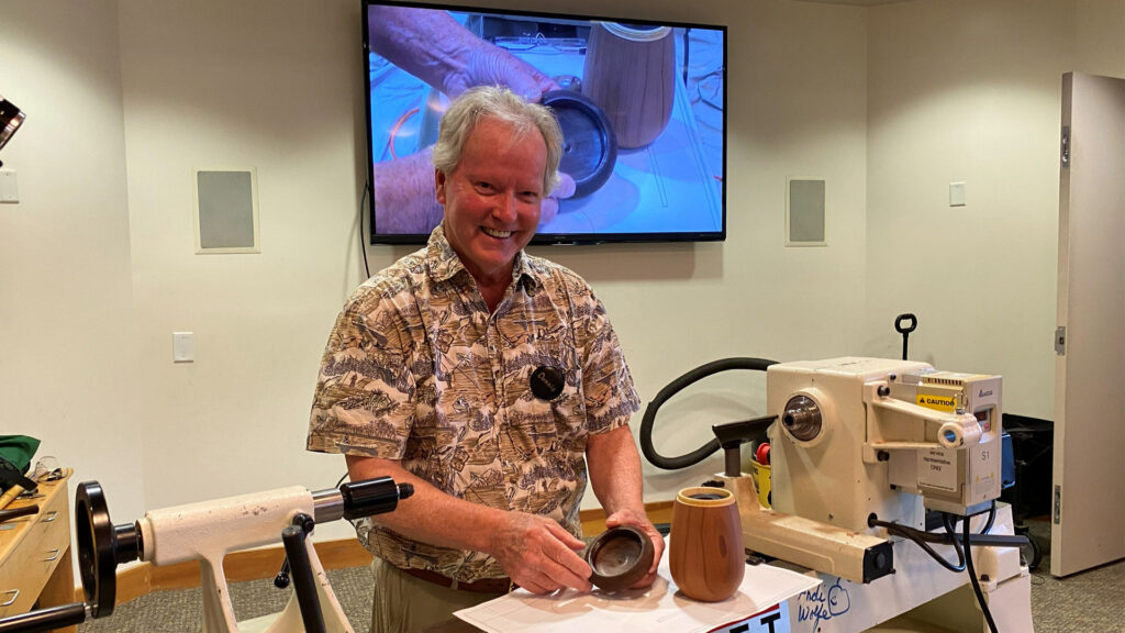 A smiling man named Dennis, identified by his name tag as Dennis Lillis, stands behind a white wood lathe at a West Bay Woodturners club meeting. He holds a small, dark wooden bowl in his hands while a larger, light-brown turned vessel sits on the machine bed beside him. Behind him, a large wall-mounted television displays a woodworking demonstration.