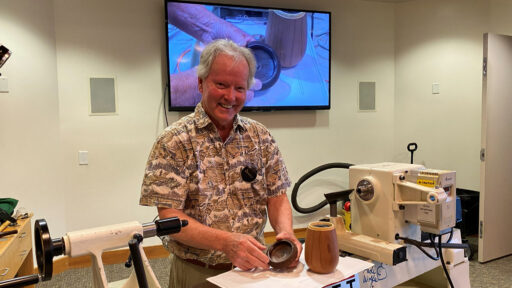 A smiling man named Dennis, identified by his name tag as Dennis Lillis, stands behind a white wood lathe at a West Bay Woodturners club meeting. He holds a small, dark wooden bowl in his hands while a larger, light-brown turned vessel sits on the machine bed beside him. Behind him, a large wall-mounted television displays a woodworking demonstration.
