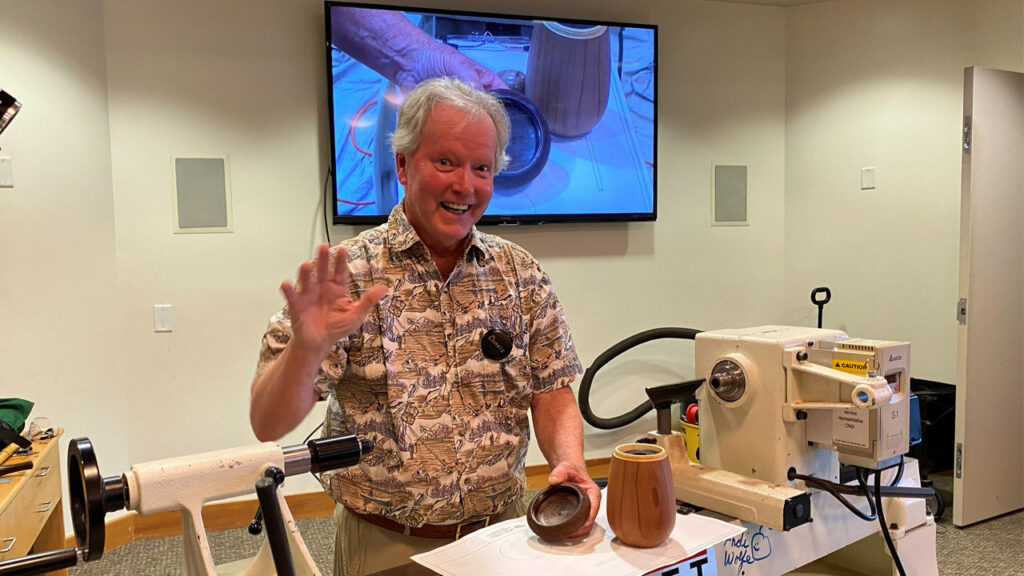 At a West Bay Woodturners meeting, a smiling man with white hair stands behind a large white wood lathe, waving to the camera with his right hand. He holds a small dark wooden bowl in his left hand, while a taller, light-colored wooden cup sits on the machine bed next to it. Behind him, a large television screen mounted on the wall displays a video close-up of a woodturning demonstration.