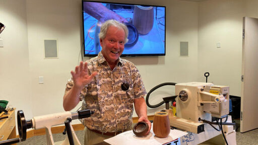 At a West Bay Woodturners meeting, a smiling man, Dennis Lillis, with white hair, stands behind a large white wood lathe, waving to the camera with his right hand. He holds a small dark wooden bowl in his left hand, while a taller, light-colored wooden cup sits on the machine bed next to it. Behind him, a large television screen mounted on the wall displays a video close-up of a woodturning demonstration.