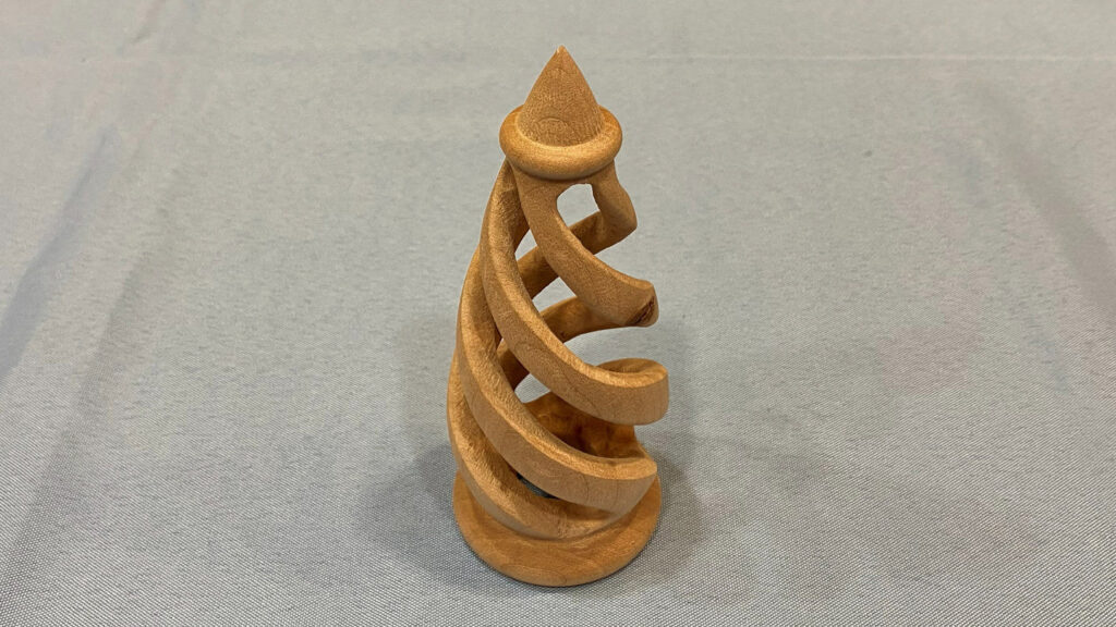 A light-colored wooden sculpture featuring a continuous, twisting spiral design that winds upward from a flat circular base to a pointed conical top. The piece appears to be a turned-and-carved wood project displayed on a grey cloth surface.