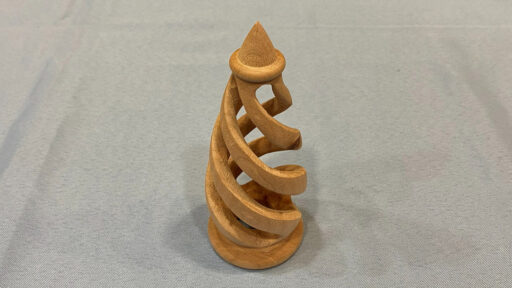 A light-colored wooden sculpture featuring a continuous, twisting spiral design that winds upward from a flat circular base to a pointed conical top. The piece appears to be a turned-and-carved wood project displayed on a grey cloth surface.