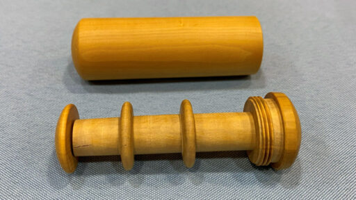 Two hand-turned wooden objects rest on grey fabric. The top piece is a smooth, light-brown hollow cylinder with rounded ends. Below it lies its matching spindle, featuring two raised flanges along the shaft, screw threads at one end, and a wider cap designed to fit into the cylinder above. Together they form a threaded container for storing sewing threads used in clothing mending.