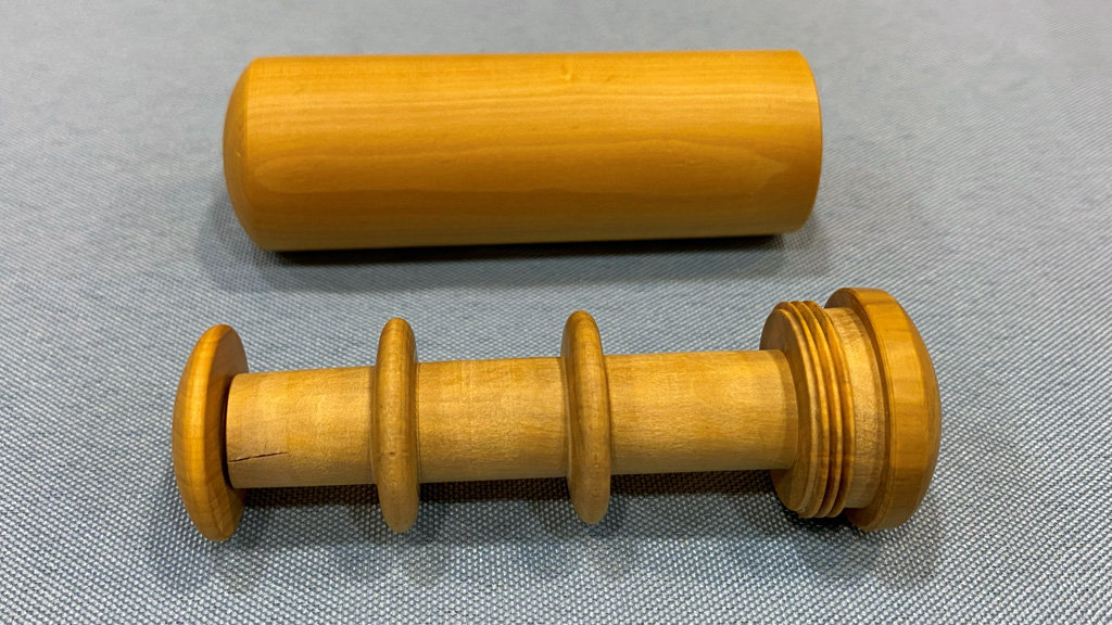 Two hand-turned wooden objects rest on grey fabric. The top piece is a smooth, light-brown hollow cylinder with rounded ends. Below it lies its matching spindle, featuring two raised flanges along the shaft, screw threads at one end, and a wider cap designed to fit into the cylinder above. Together they form a threaded container for storing sewing threads used in clothing mending.