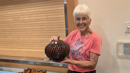 Dottie Bang smiles while holding a dark brown wooden vessel that she turned and carved slats arranged in a spiral pattern. She is wearing a pink T-shirt with a floral design. The image was taken at the West Bay Woodturners meeting, where members displayed their woodturning projects.