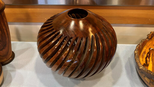 A glossy, dark brown wooden vessel with a bulbous, spherical shape featuring deep, spiraling carved grooves that wrap around its surface. The piece has a high-shine finish and an opening at the top revealing inner vertical ribs. It sits on a light-blue cloth table cover, flanked by partial views of other wooden bowls, displayed as part of a woodturning project showcase.