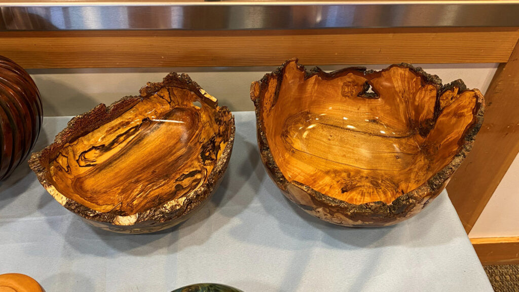 Two finished live-edge wooden bowls sitting side-by-side on a light blue display cloth. The bowl on the left features rich, dark amber grain with dramatic black swirls and streaks, while the bowl on the right is a lighter golden-orange hue with smoother figuring. Both bowls have been turned to a glossy finish but retain their rough, natural bark edges around the rim.