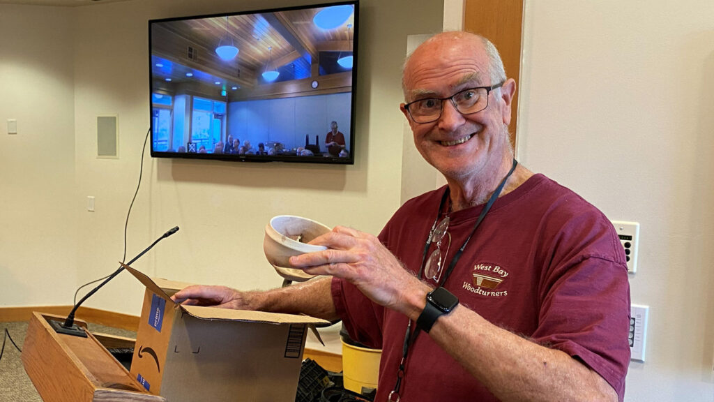 A smiling man, Edgar Whipple, with glasses and a maroon t-shirt featuring the "West Bay Woodturners" logo, stands next to a wooden podium, holding up a light-colored wooden bowl in his left hand while resting his right hand on an open cardboard box. Behind him, a large wall-mounted TV screen shows a live feed of the room's interior and audience members seated under blue lighting. The photo shows the WBW club meeting in June 2026.