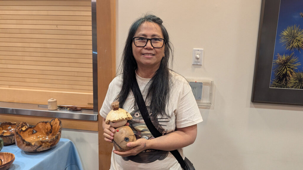 Edy Chung, a woman with long dark hair and black-rimmed glasses, stands indoors, holding a small wooden turning project resembling a figure with textured hair and painted facial features. She is wearing a white T-shirt featuring a Totoro graphic and has a black strap across her shoulder. To her left, several large, polished wooden bowls are displayed on a table covered with a light blue cloth, sitting in front of a window with closed blinds. To her right, a framed picture of yellow plants hangs on the wall. The image captures a moment from the West Bay Woodturners meeting where members display their projects.