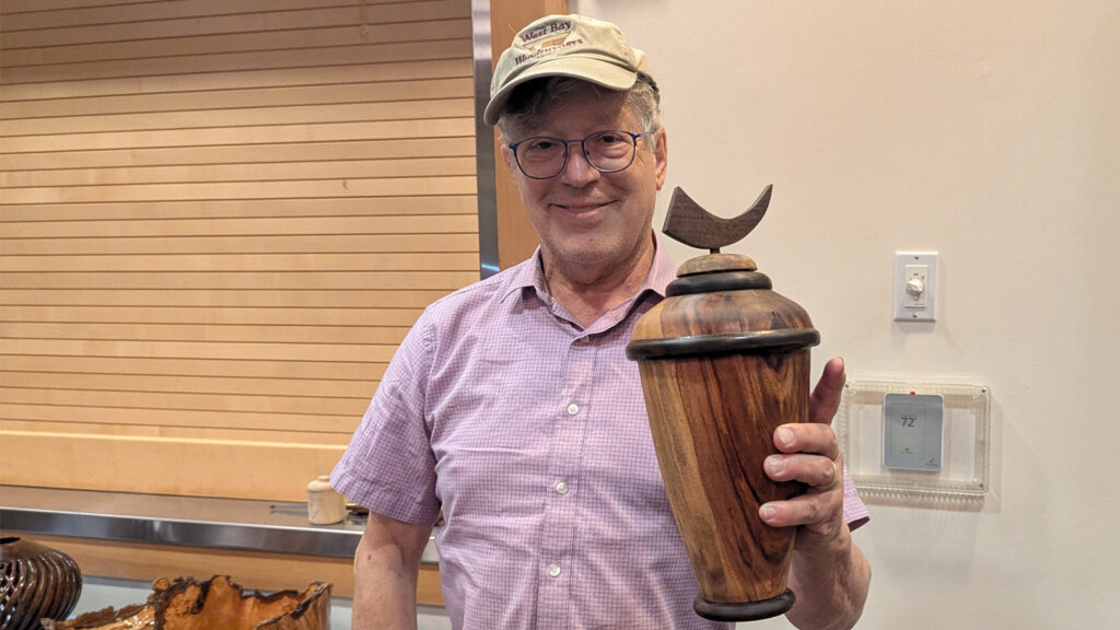 Fred Colman smiles while holding up a turned camphor wood vase topped with a crescent moon finial. He is wearing glasses, a pink checkered shirt, and a beige cap that reads "West Bay Woodturners," standing in front of a light wooden slat wall with other wood projects visible on a table to his left.